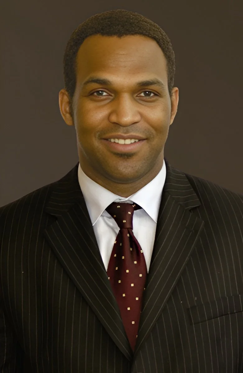 Portrait of a man in a business suit with a white shirt and burgundy tie, smiling, against a dark background.