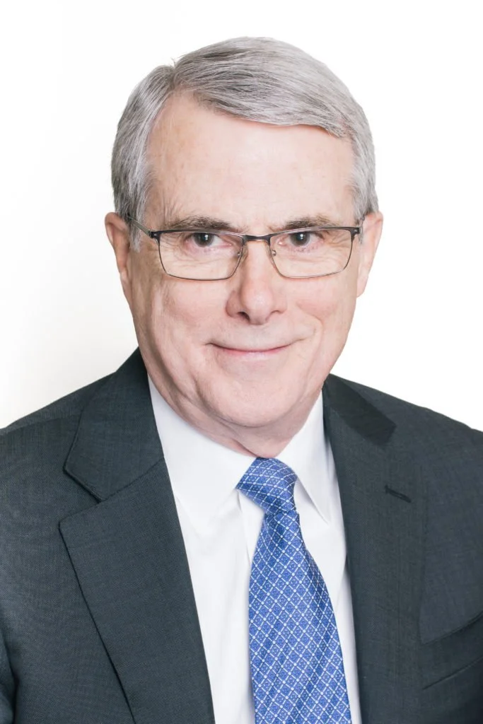 Headshot of an older man with glasses, wearing a dark suit, white shirt, and blue patterned tie, smiling against a white background.