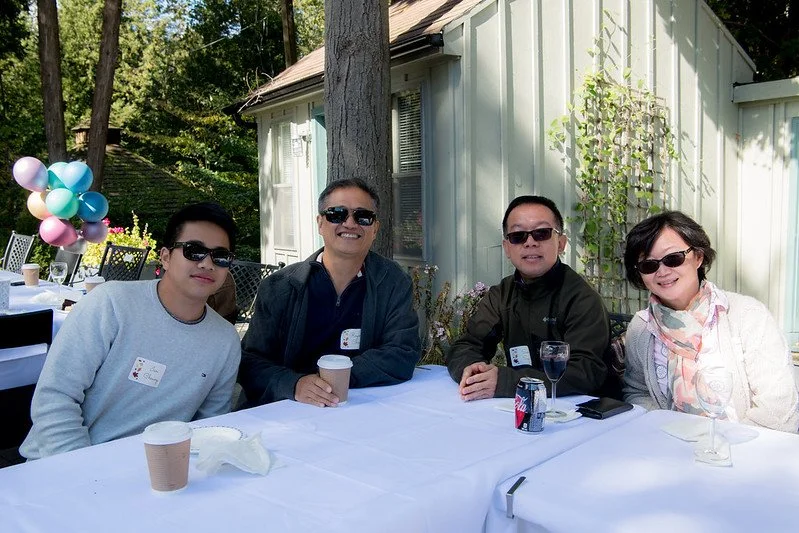 Four people sitting at a table outdoors during daytime, smiling, with balloons in the background, a tree, and a small house or shed. The table has drinks and napkins.