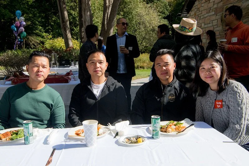 Group of four people sitting at an outdoor table with food, surrounded by other people standing and socializing in a park-like setting with trees.