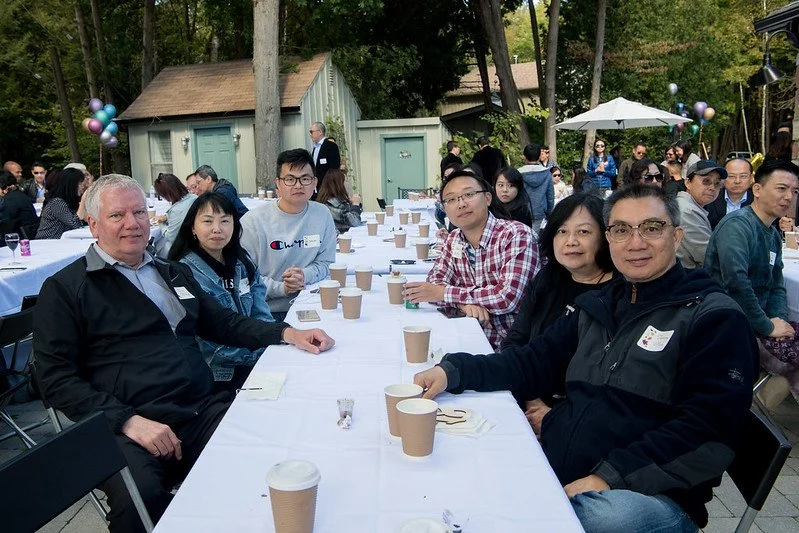 People sitting at a long outdoor table during a gathering or celebration, with cups and plates on the table, trees in the background, and balloons and a shed visible.