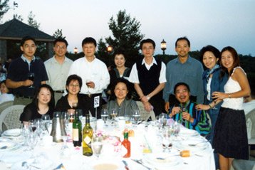 Group of people gathered around a table outdoors during evening, with some seated and some standing, celebrating at a social event.