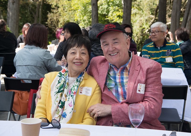 Two smiling people, a woman and a man, seated at an outdoor event among other attendees. The woman wears a yellow blazer and a floral scarf, while the man wears a pink blazer over a plaid shirt and a green bow tie. There are trees in the background.