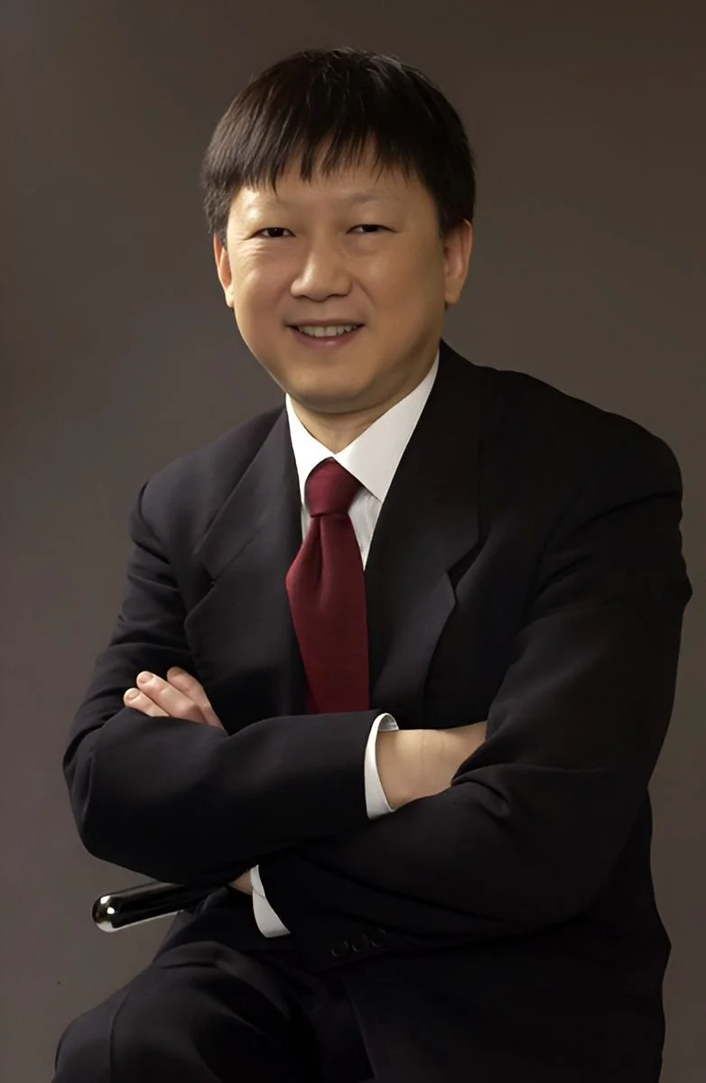 Professional portrait of an Asian man in a black suit with white shirt and maroon tie, smiling with arms crossed, against a dark background.