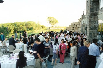 Outdoor event with round tables and chairs, many people socializing and standing around, historic stone building in the background, green trees and a clear sky.
