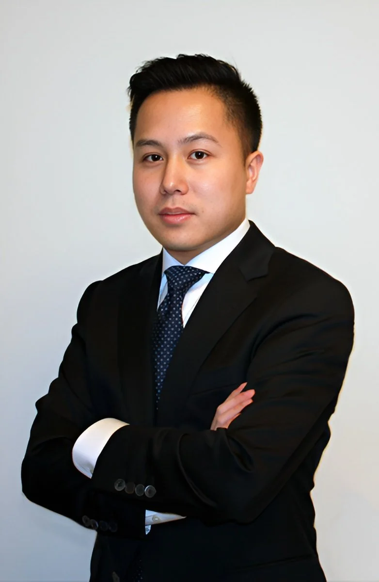 Professional man in a black suit and blue tie, arms crossed, against a plain white background.