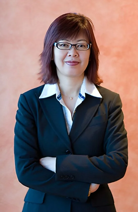 A woman with shoulder-length dark hair, wearing glasses, a white collared shirt, and a dark blazer, standing with her arms crossed in front of a pinkish-orange background.