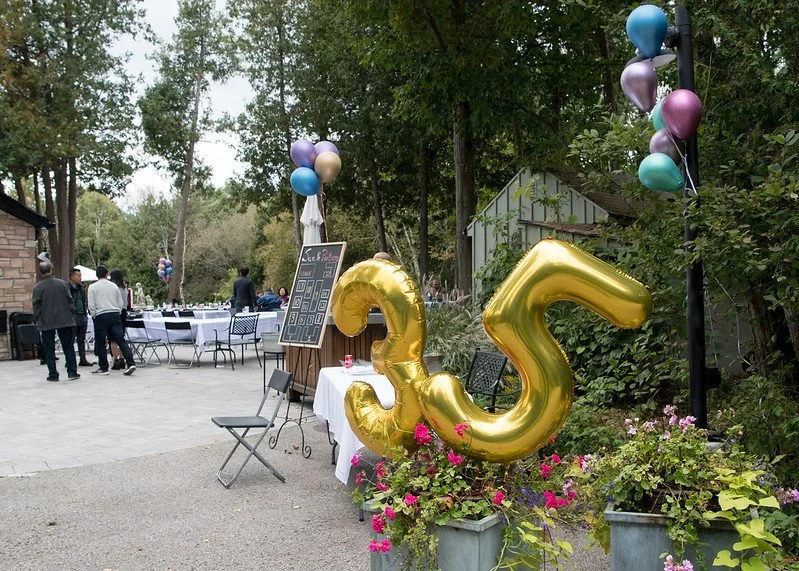 Decorative 3rd birthday celebration with large gold number 3 balloon, colorful balloons, tables with white tablecloths, and guests in an outdoor garden setting.