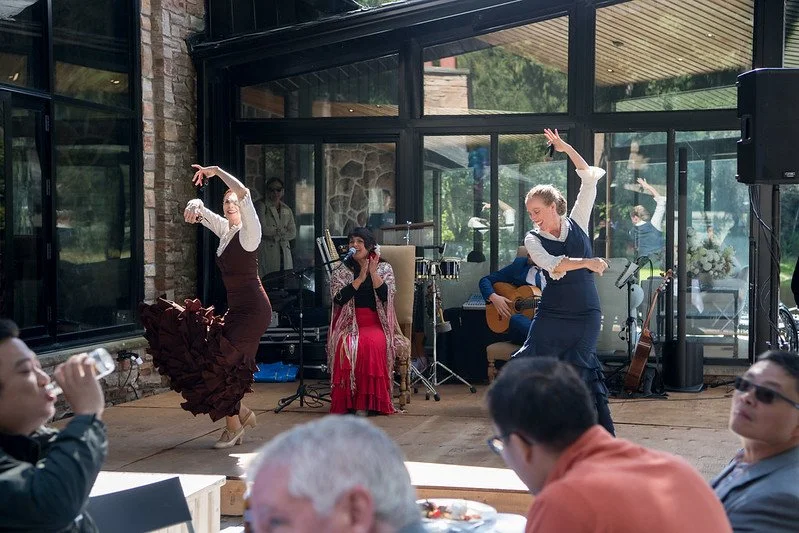 Dancers performing flamenco dance on a wooden floor, with musicians playing guitar and singing in the background, at a lively indoor event with guests watching.