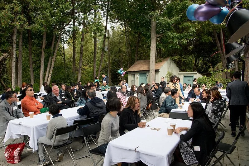 People gathered at outdoor event seated at long tables with white tablecloths, surrounded by trees and a small building, with balloons and outdoor lighting.