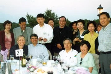 A group of people at a festive outdoor dinner gathering, smiling and posing for the photo with a table of food and drinks in front of them.