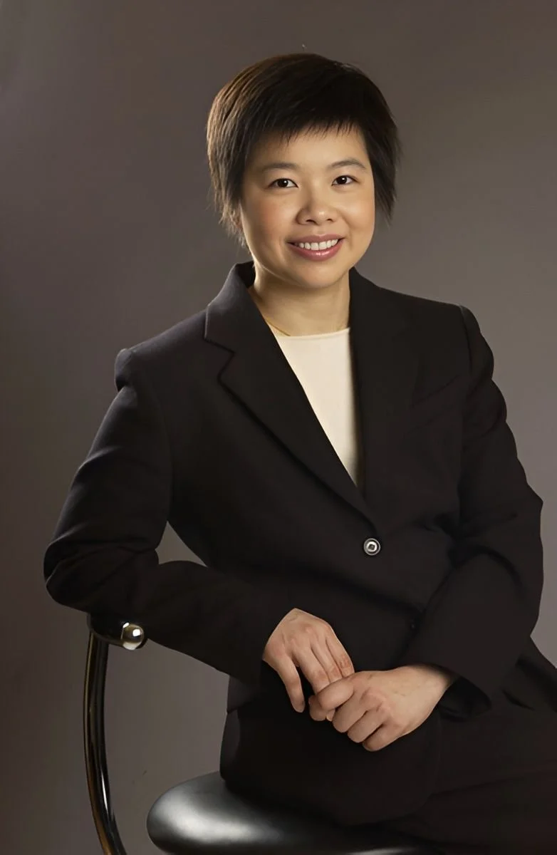 A woman with short black hair wearing a black blazer and cream blouse, sitting on a black chair against a gray background, smiling at the camera.