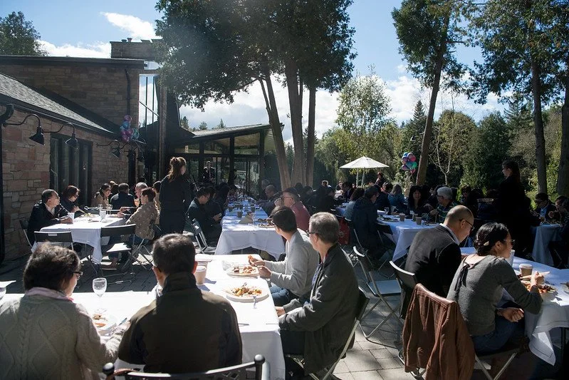 Outdoor gathering of people seated at tables with white tablecloths, enjoying a meal on a sunny day surrounded by trees and a building.