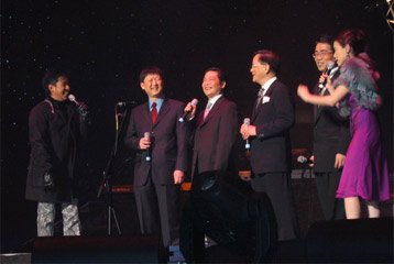 Group of five men and one woman standing on a stage, smiling and talking, with one of the men holding a microphone.