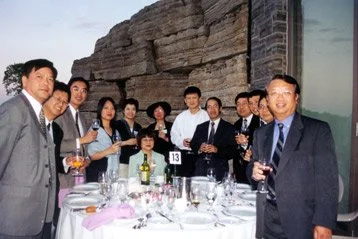 Group of people dressed in formal attire at a dinner party, holding glasses of wine, around a table with drinks and tableware, in a well-lit indoor setting.