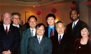 Group of eight people dressed in formal attire, posing for a photo at an indoor event with red hanging decorations.