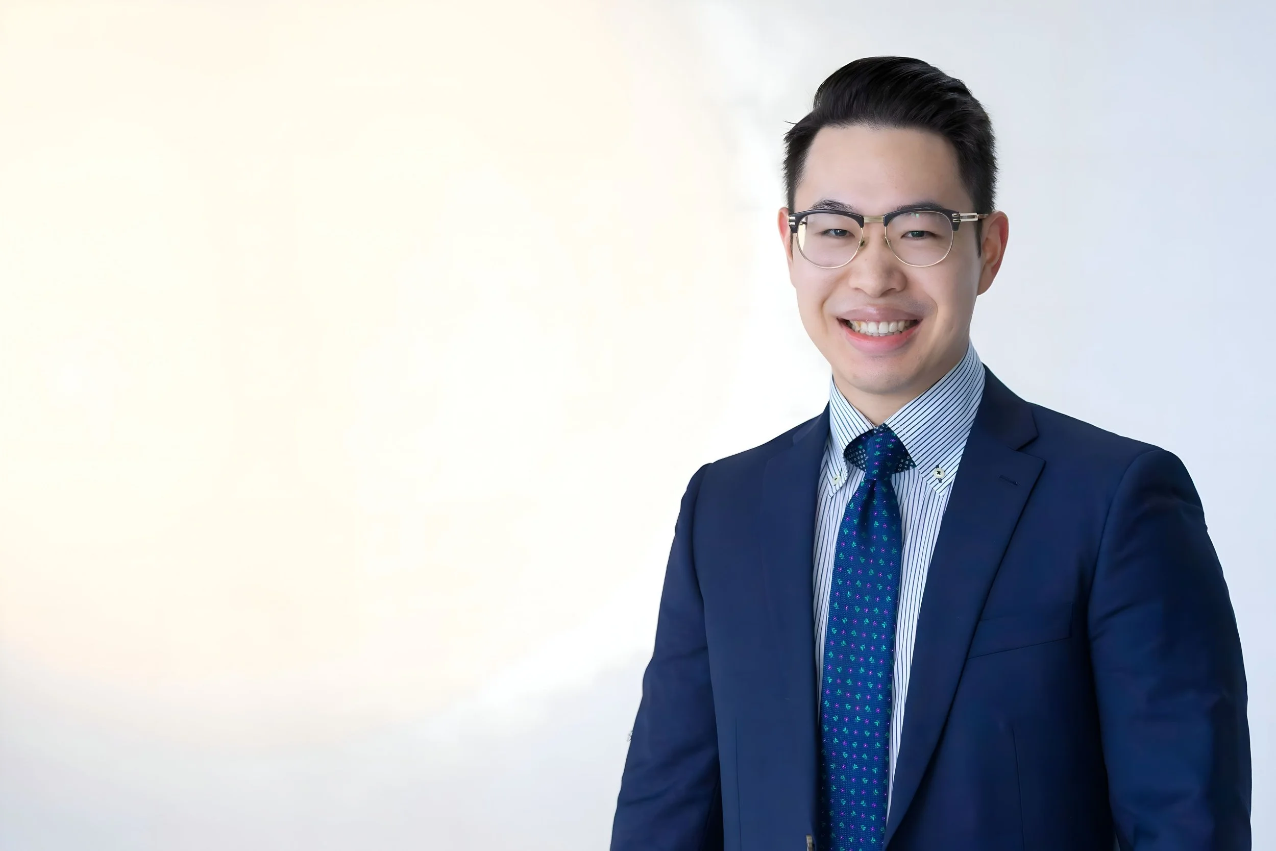 Smiling Asian man wearing glasses, a navy suit, a striped shirt, and a patterned tie, standing against a light background.