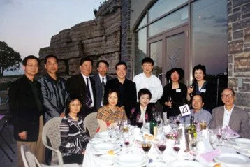 A group of 11 people gathered around a dining table outdoors, with some standing and others sitting, during an evening meal with wine glasses and table settings, in front of a brick building and a large glass window.