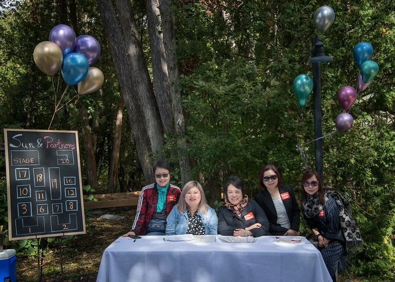 Five women sitting at a registration table outdoors with a blackboard sign and colorful balloons, surrounded by trees.