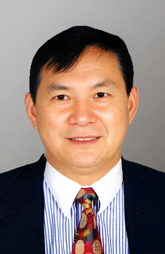 A portrait of an Asian man in business attire, wearing a dark suit, a white shirt with blue stripes, and a multicolored tie, smiling against a gray background.