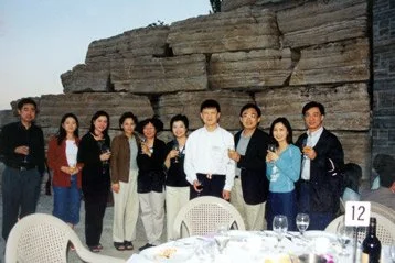Group of people standing together outdoors near a large rock formation, with a table set for dining in the foreground.