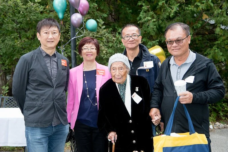 Group of five people standing outdoors with balloons and greenery in the background, including an elderly woman in the center holding a cane and wearing a hat, surrounded by four adults.
