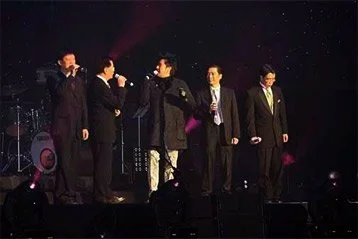 Five men dressed in suits and a coat on stage with a dark background and lights, possibly at a formal event or performance.