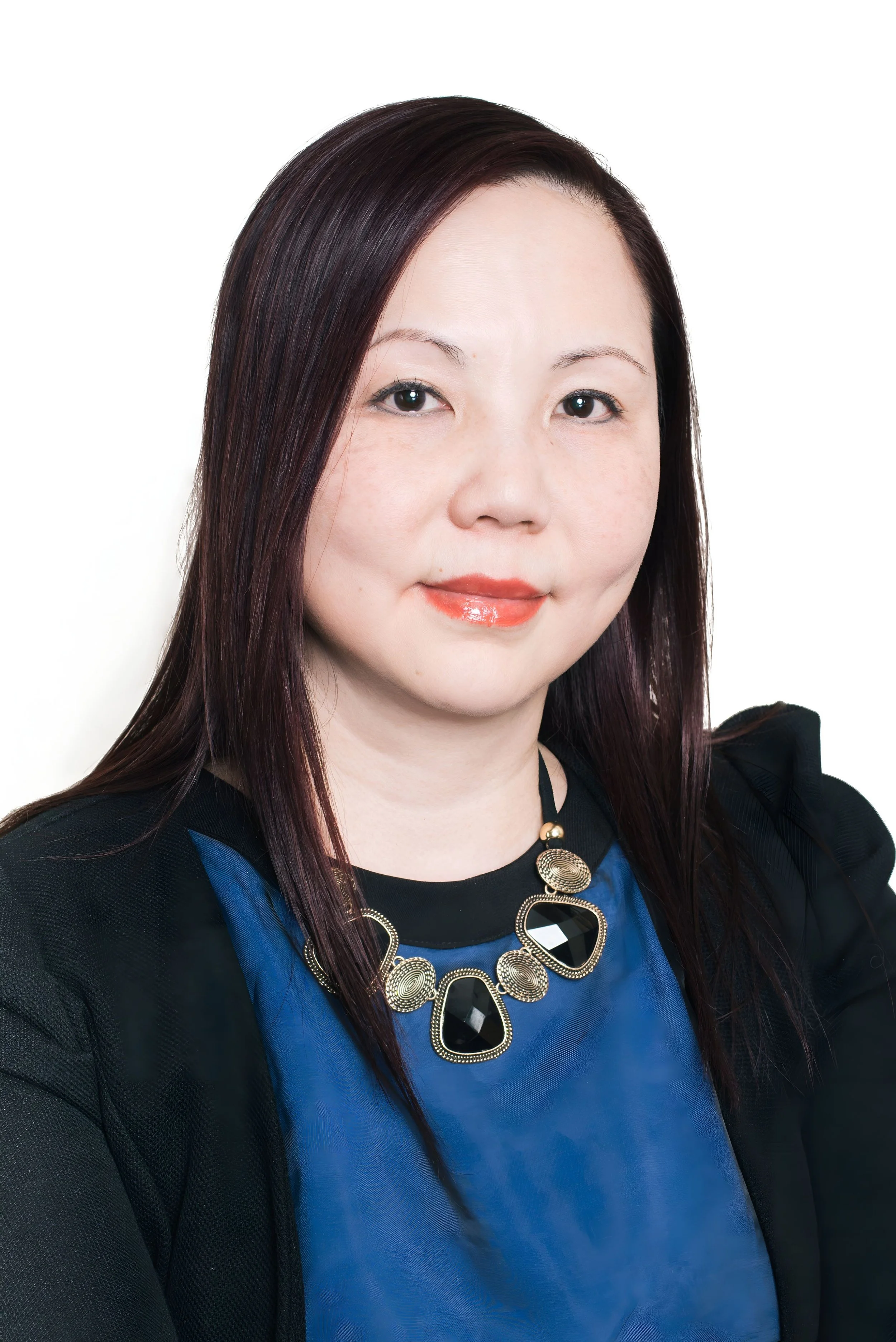 A woman with long dark hair, wearing a blue top, black jacket, and a statement necklace, smiling softly against a plain white background.