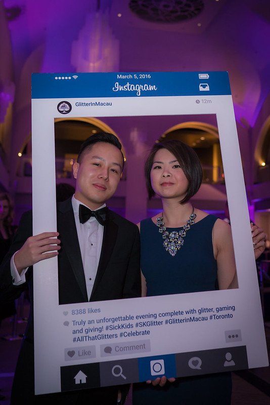 Two well-dressed individuals, a man in a tuxedo and a woman in a blue dress with a statement necklace, holding a large pretend Instagram photo frame at a festive event with purple lighting and arched ceilings.