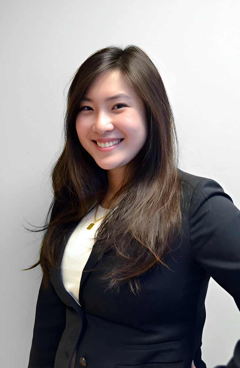 A young woman with long brown hair smiling and wearing a black blazer over a white top, standing against a plain gray wall.