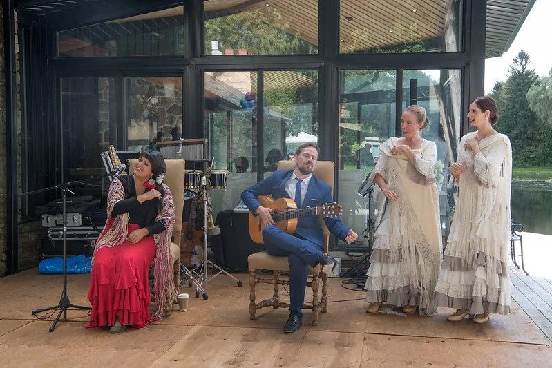 Four people, two women dressed in vintage style cream dresses and two performers, one woman in a long red skirt and black top and a man in a blue suit playing a guitar, are on a wooden stage near a glass building with a pond and trees in the background.