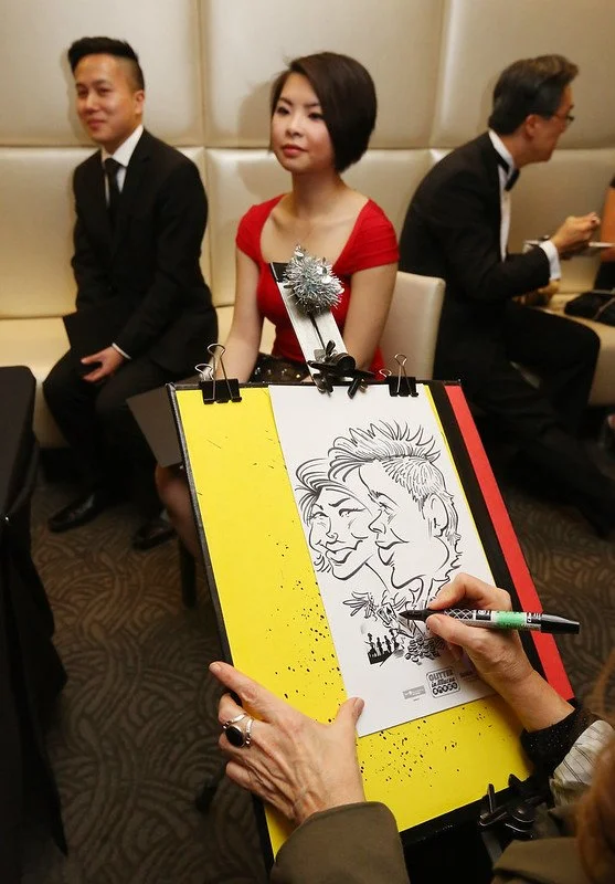 A caricature artist drawing a cartoon of a woman with short hair and a man with spiky hair at a formal event, with two seated guests in the background, one in a red dress and the other in a black suit.