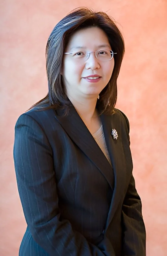 Professional woman in a dark pinstriped blazer and glasses, standing against a peach-colored background.