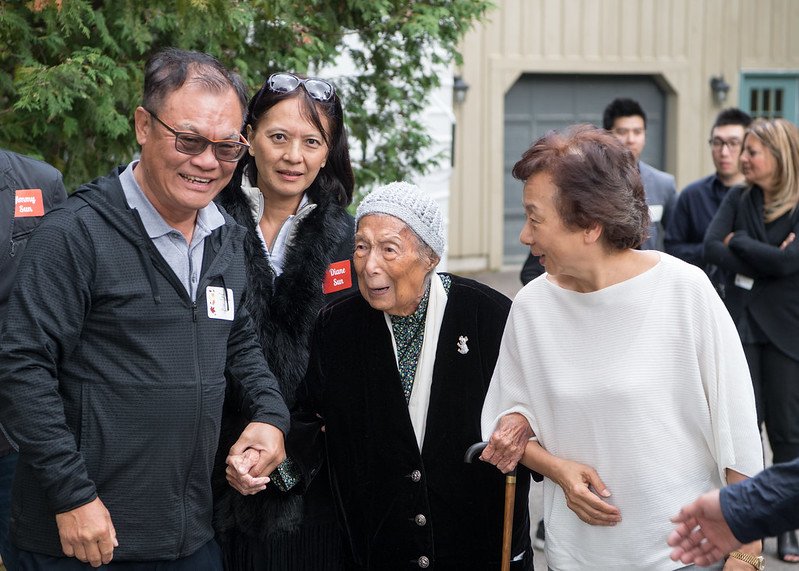 A group of five people, including elderly women and middle-aged men and women, gathered outdoors in a yard or garden area, smiling and talking with each other.