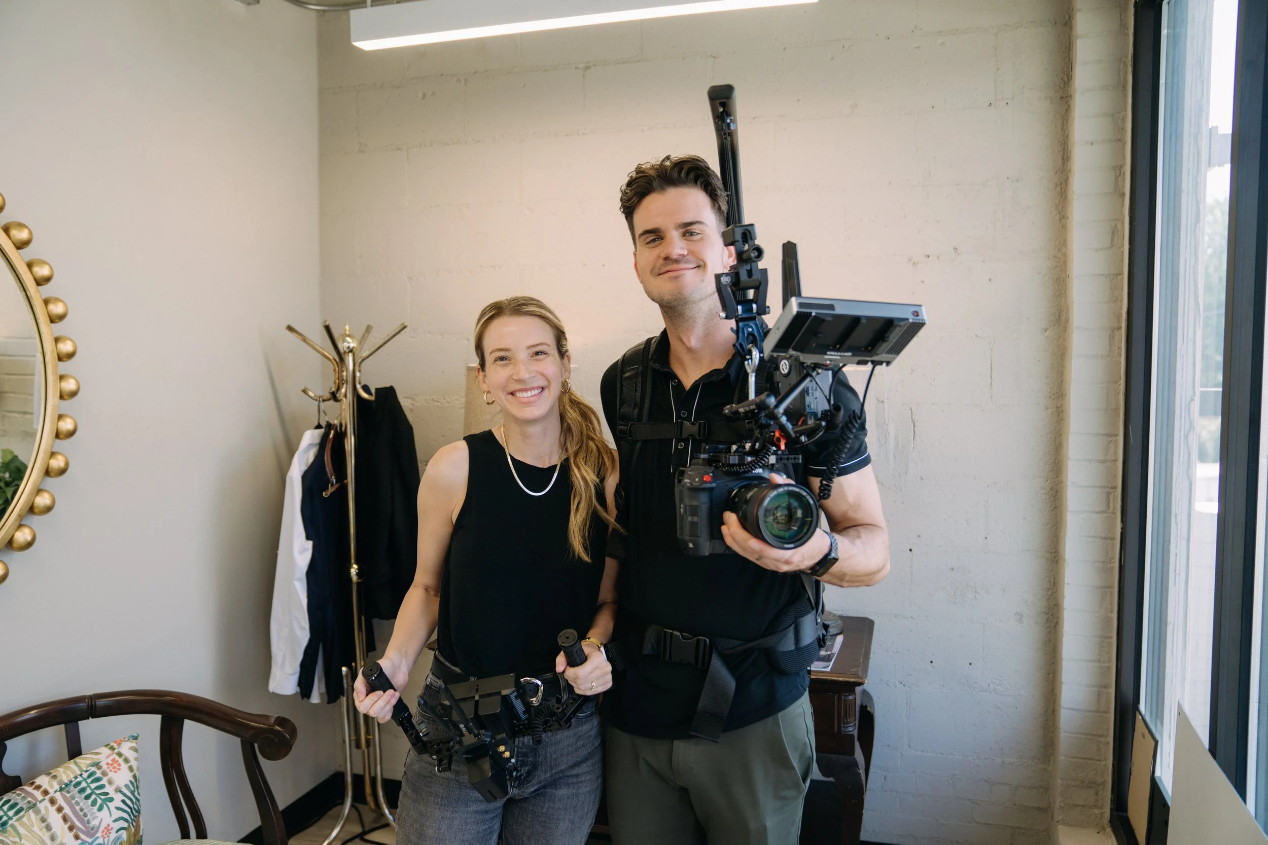 Two smiling people, a woman and a man, posing with camera equipment in a brightly lit room with a mirror, coat rack, and window.