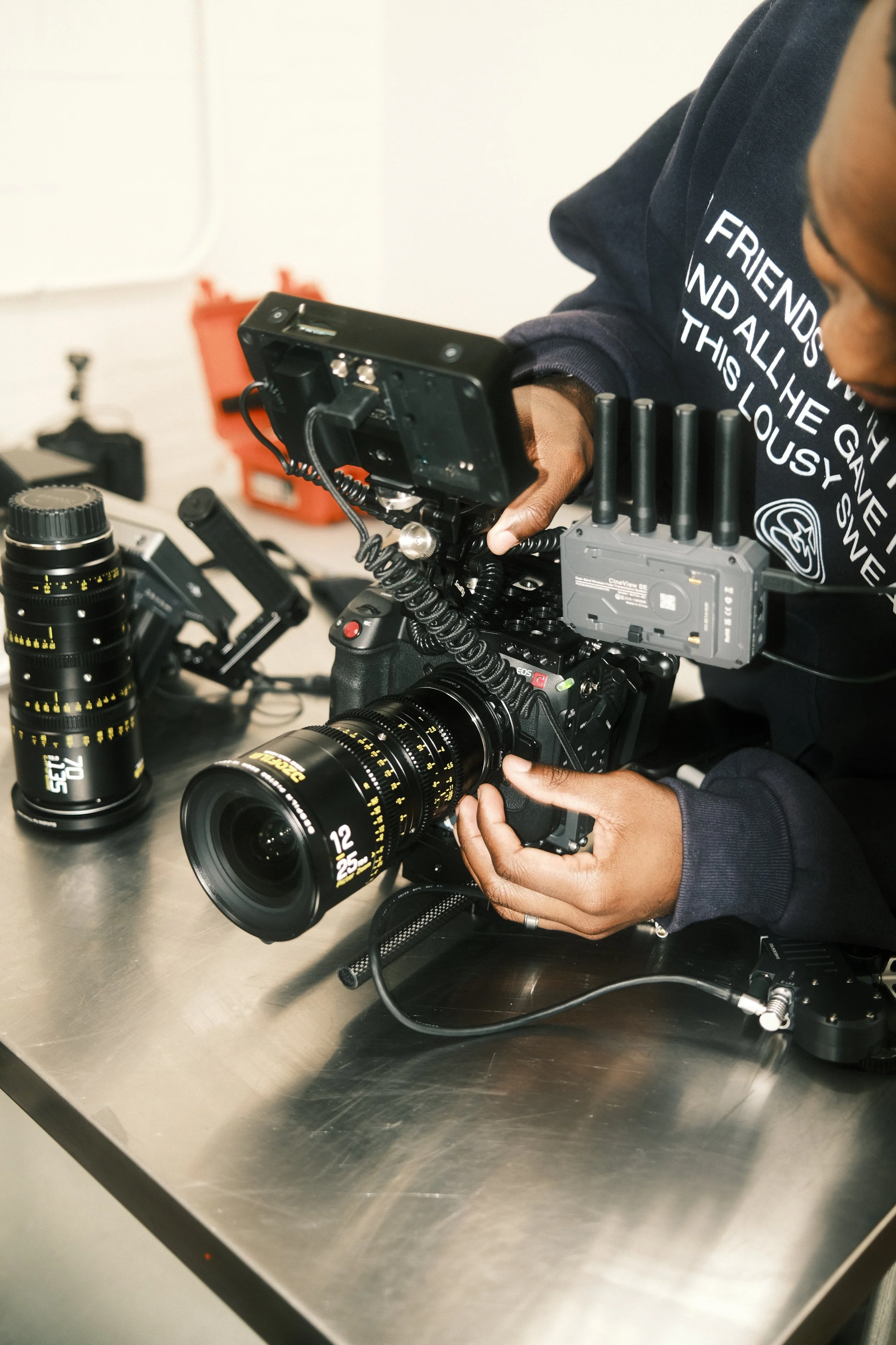 Person operating a professional video camera with a mounted external monitor and multiple lenses on a metal table.