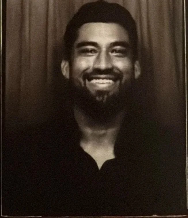 Black and white photo of a smiling man with short dark hair, a beard, wearing a dark top, posing in front of a wooden background. photo strip print from luna & light photo booth. Bay Area California