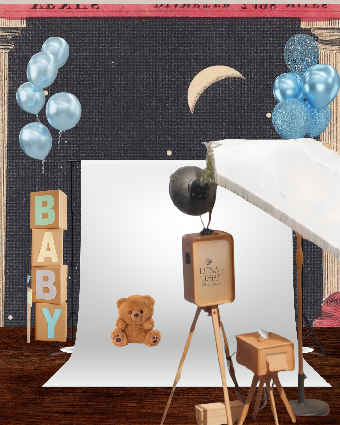 Baby photography setup with a white backdrop featuring a teddy bear, decorative blue balloons, a mount of blocks spelling 'BABY', and lighting equipment, with a starry night moon/sky mural in the background.