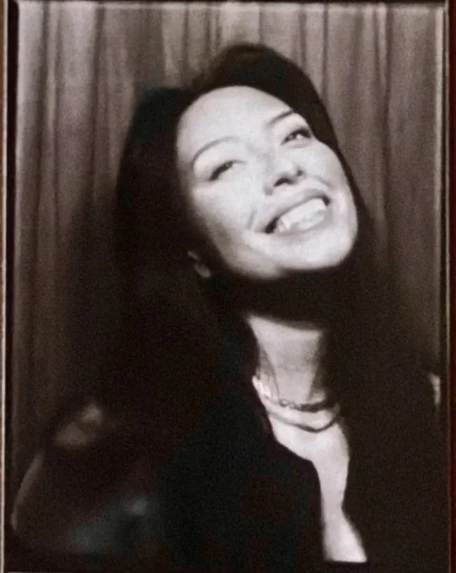 Black and white photo of a smiling woman with long dark hair, posing in front of a wooden background.