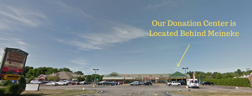 A shopping plaza with a parking lot in front, several cars parked, and a signboard on the left. Text overlay indicates the donation center is behind the Meineke. Clear sky with some clouds.
