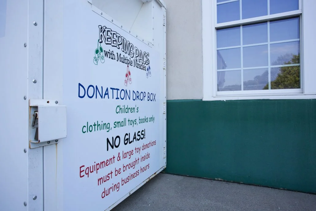 Donation drop box outside a building with a sign that reads, "Keeping hope with multiple miracles," and specifies the accepted items as children's clothing, small toys, and books, with a note that no glass or large toy donations are allowed.
