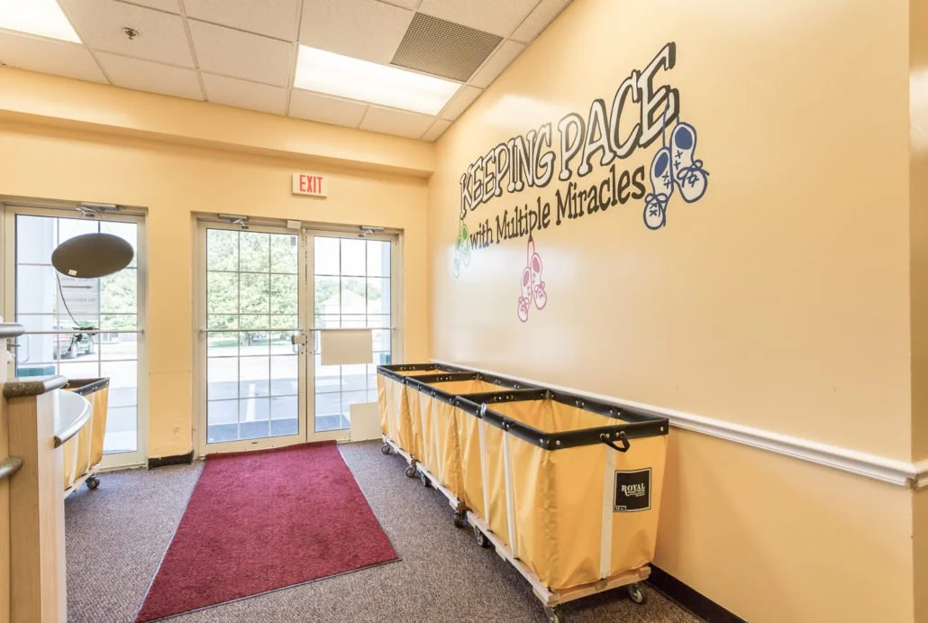 Interior of a room with yellow walls, a red carpet runner, and a wall mural that reads 'Keeping Peace with Multiple Miracles' with decorative butterfly illustrations. There are yellow storage bins on wheels along the wall, glass double doors leading outside, and an exit sign above.