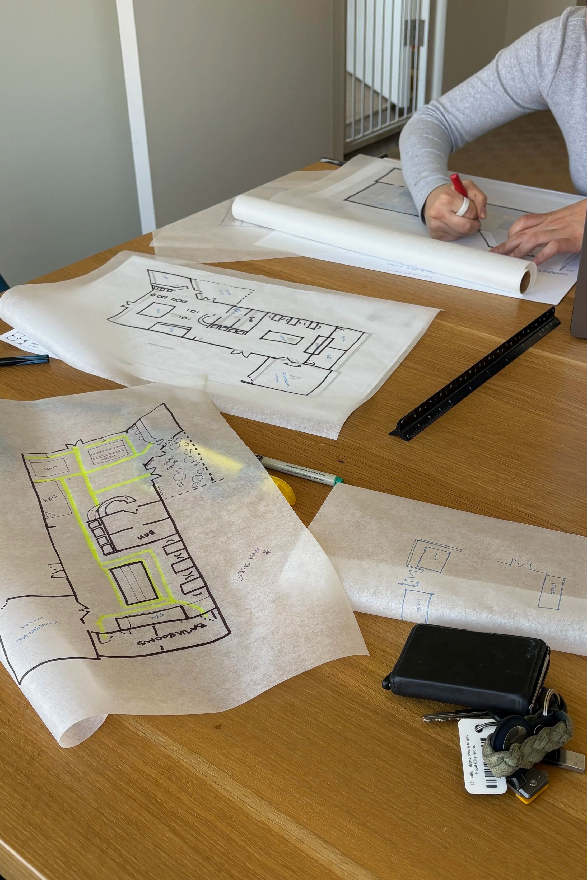 A person working on architectural blueprints spread out on a wooden table, with additional drawings, a black ruler, a smartphone, and a set of keys nearby.
