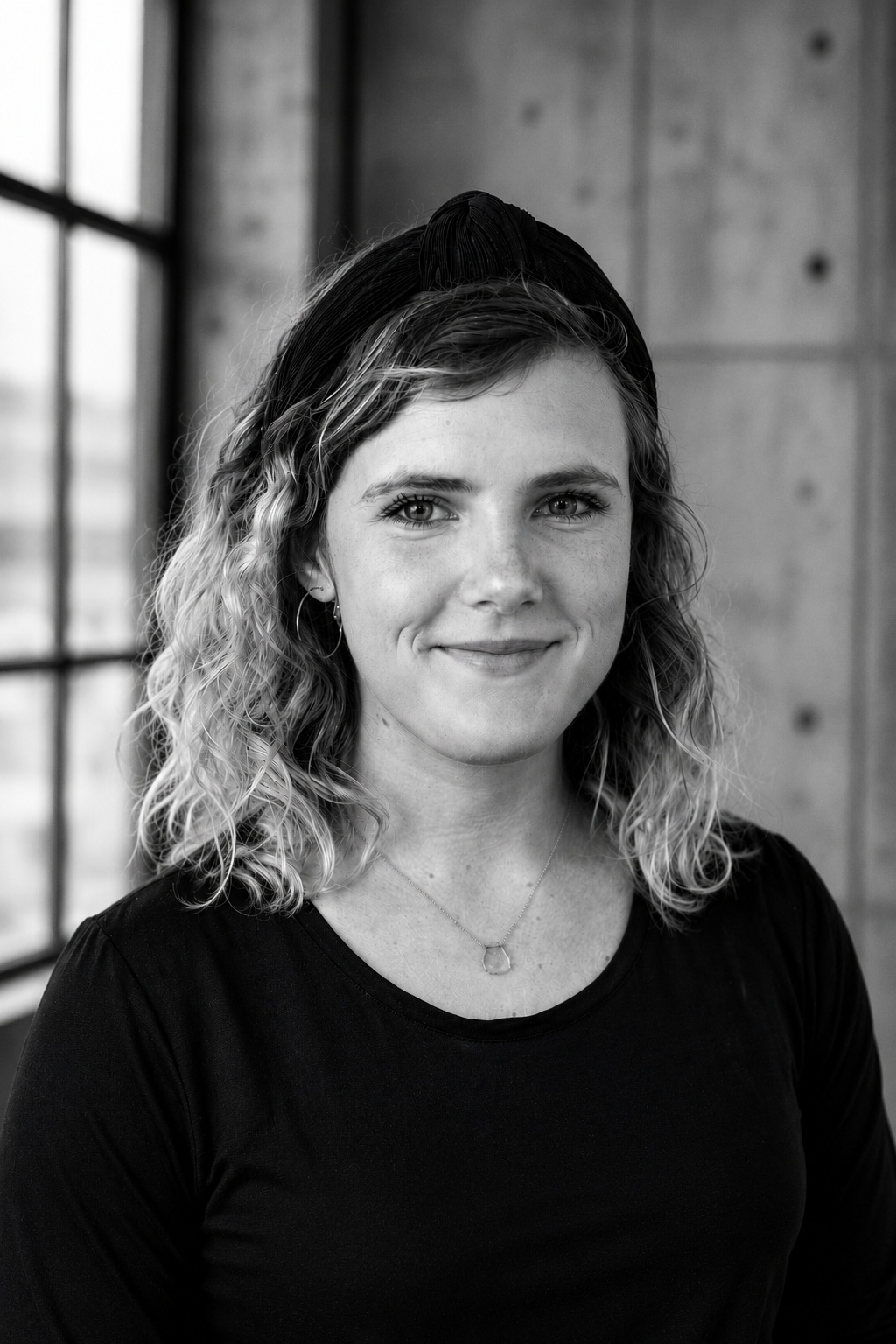 Black and white portrait of a young woman with curly hair, wearing a headband and earrings, smiling softly, standing indoors near large windows and a concrete wall.