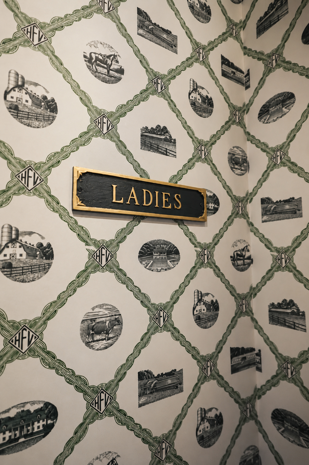 Sign labeled 'Ladies' on a wall with black and white farm and countryside illustrations connected by green decorative lines and the 'HFV' logo.