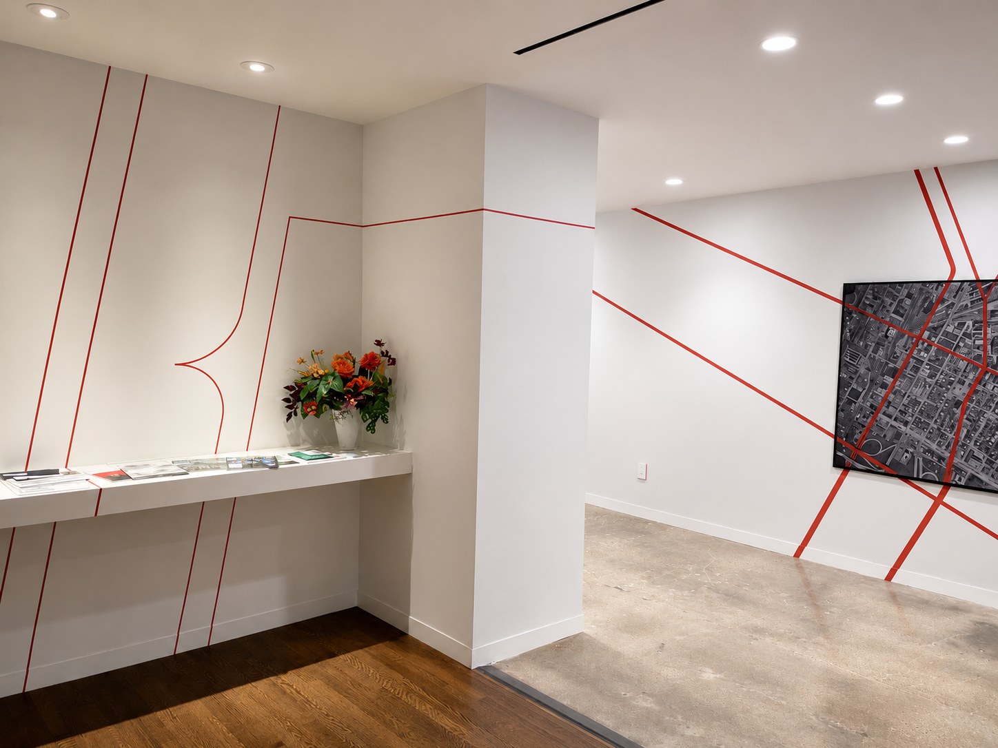 Interior of a modern gallery or office space with white walls decorated with red lines, a shelf with informational brochures and a flower arrangement, a mounted map or artwork on the wall, and recessed ceiling lights.