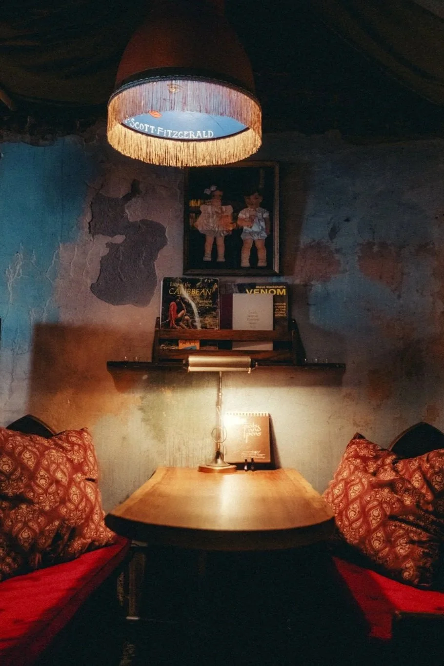 Cozy seating area in a dimly lit restaurant with a wooden table, red cushions, an overhead lampshade, and artwork on the wall.