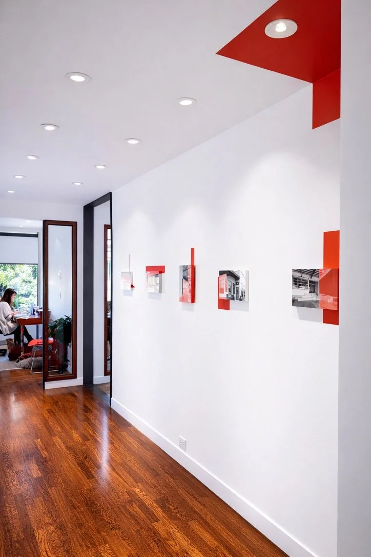A hallway in an art gallery with a white wall displaying small framed photographs with red accents. The floor is polished hardwood, and a woman is sitting at a desk in a room visible through an opening on the left side.