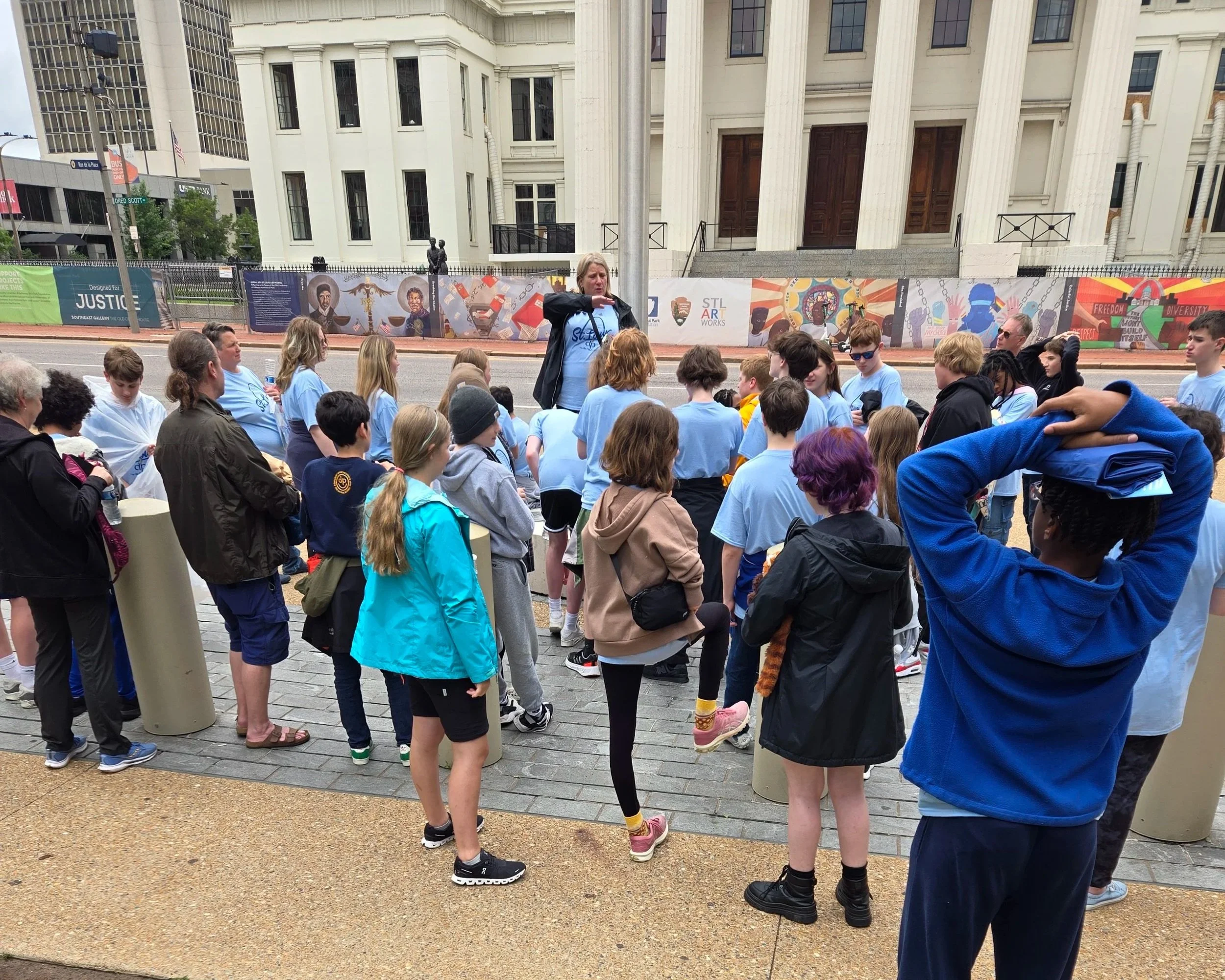 A group of people, mostly children, gathered outdoors in front of a large building with tall columns. A woman appears to be speaking to the crowd, holding a piece of paper. Behind them, there are colorful banners displayed along a fence.