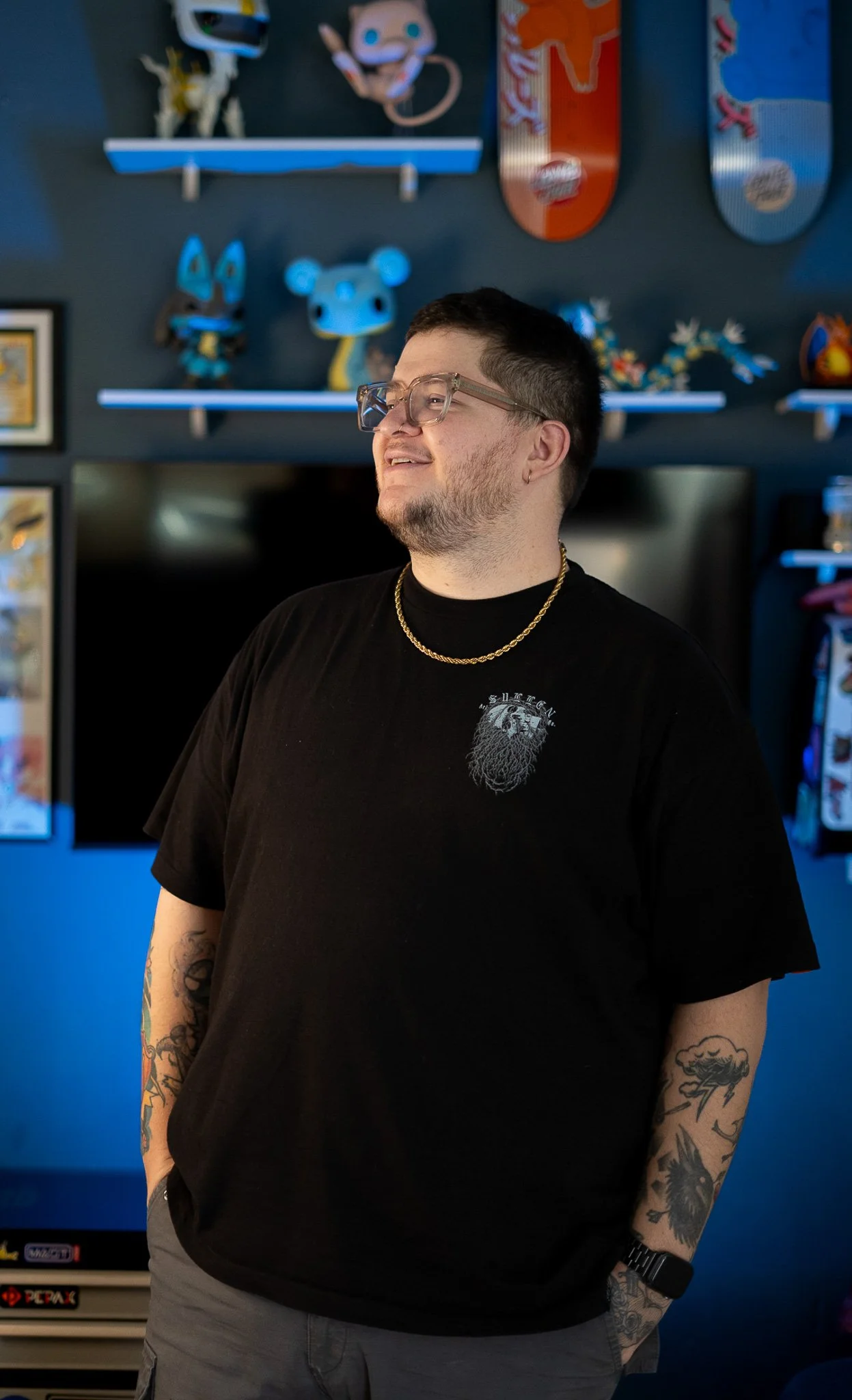 Hombre joven con gafas y barba, usando camiseta negra con un diseño en el pecho, con tatuajes en los brazos y usando reloj y collar, de pie frente a una pared decorada con figuras y objetos de decoración en tonos azules y otros colores.